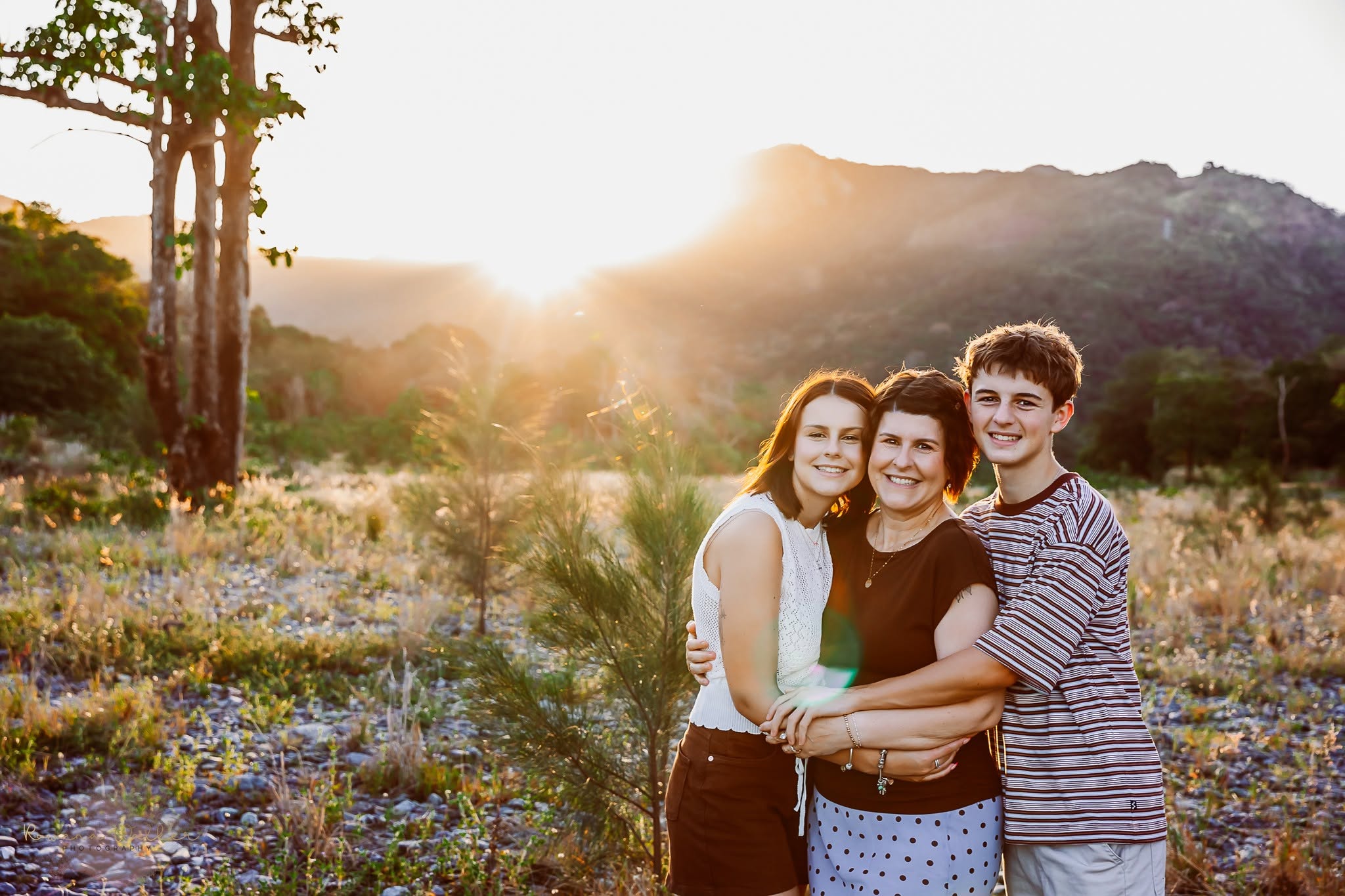 Three family members hugging in a golden field at sunset in front of a mountain