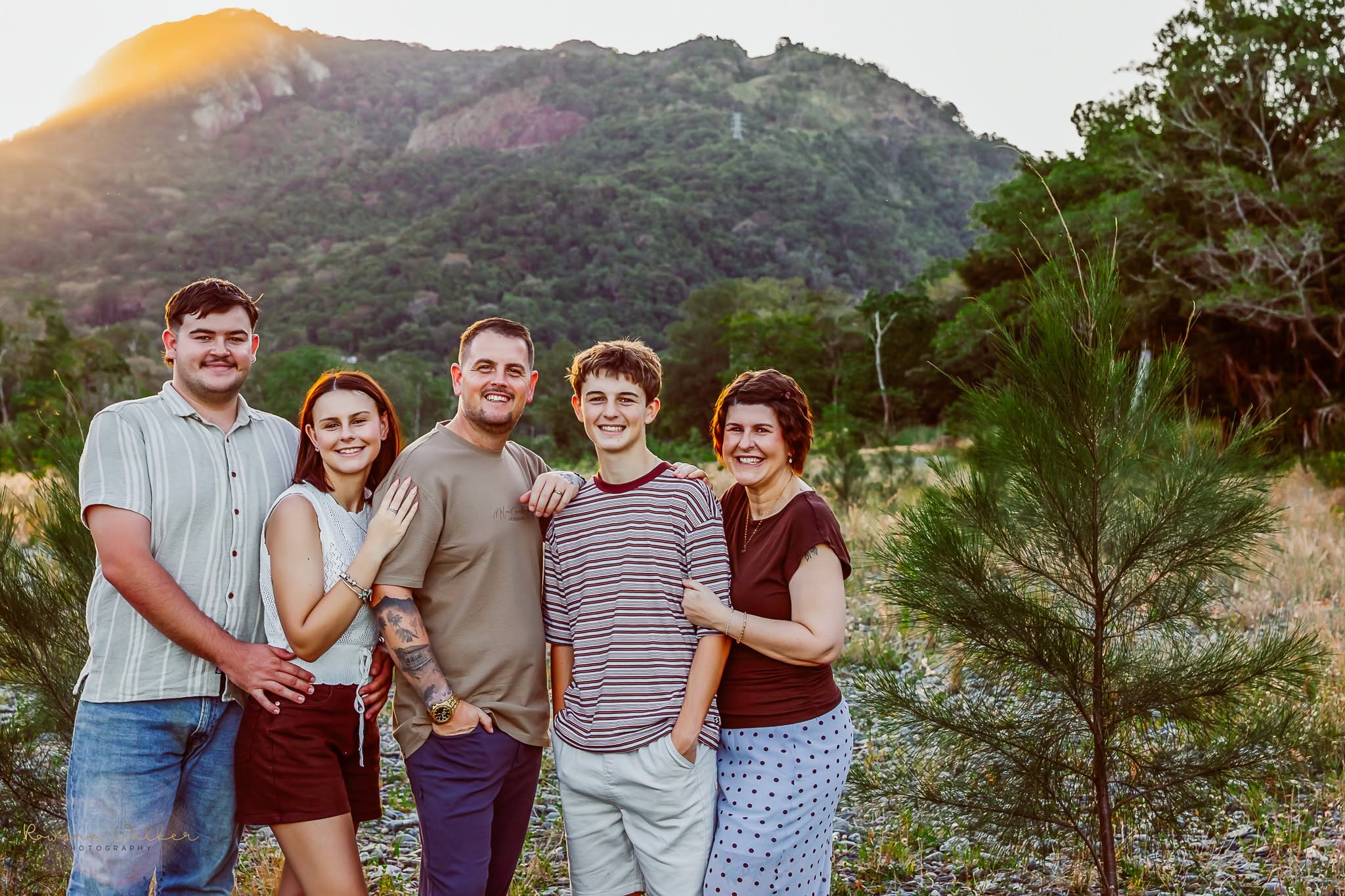 A family of five standing close together in front of a sunlit mountain