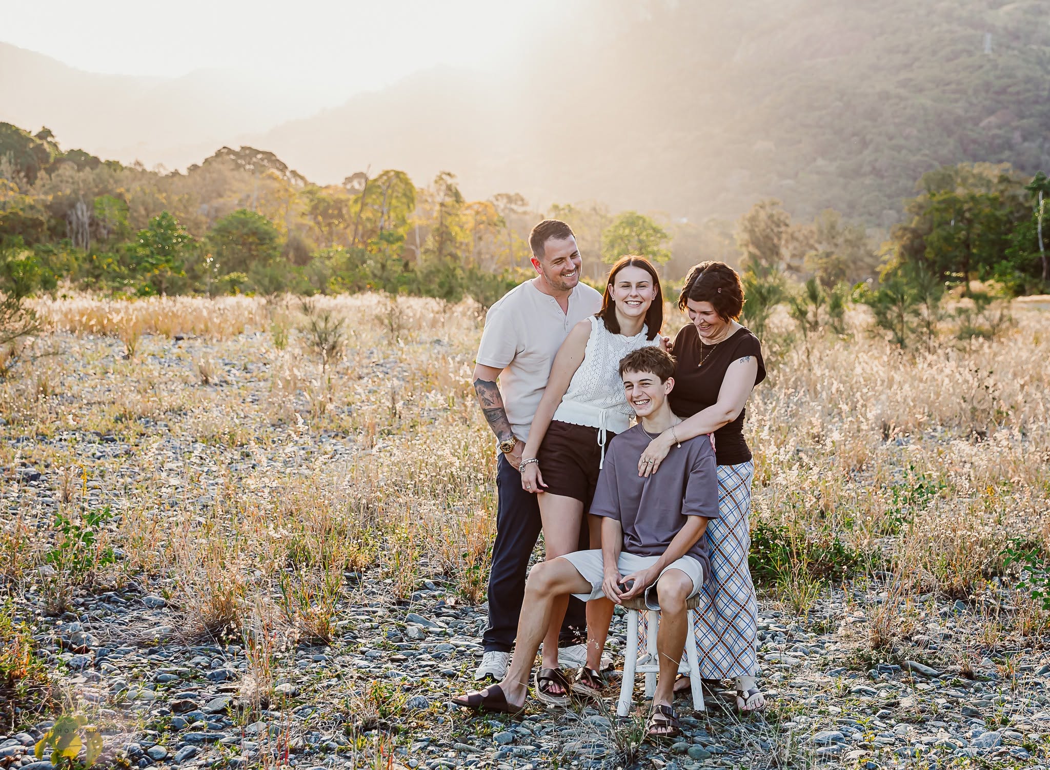 A family of four laughing together in a grassy field, one member seated on a white stool